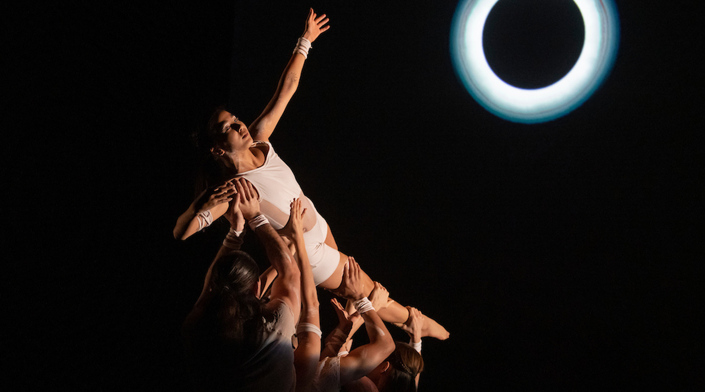 Red Sky Performance: A dancer in a white bike unitard, is held in the air on their side with one arm reaching up, by three other dancers. A ring of light that looks like a solar eclipse is on the black background.