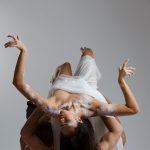 Red Sky Performance: A group of shirtless people with their backs the camera holding up a woman facing the sky. She has a white cloth wrapped around her and white body paint on her arms and shoulders. She reaches up to the sky and her hair hangs down to the ground. The background is grey.
