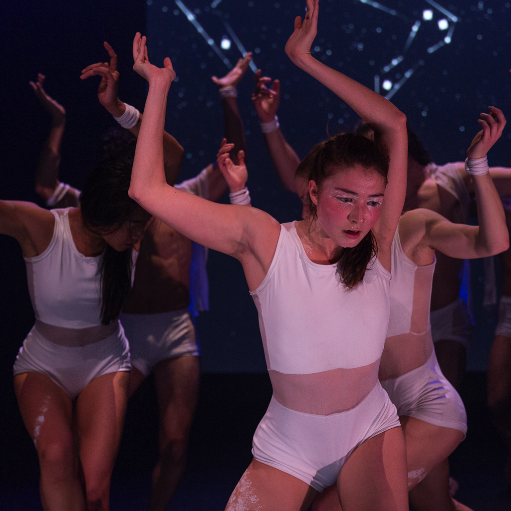 Red Sky Performance: A group of people in white leotards and shorts with mesh around their waists. They are all squatting holding their arms above their head that are bent at the elbow and wrist. They have white body paint on their eyes and thighs.