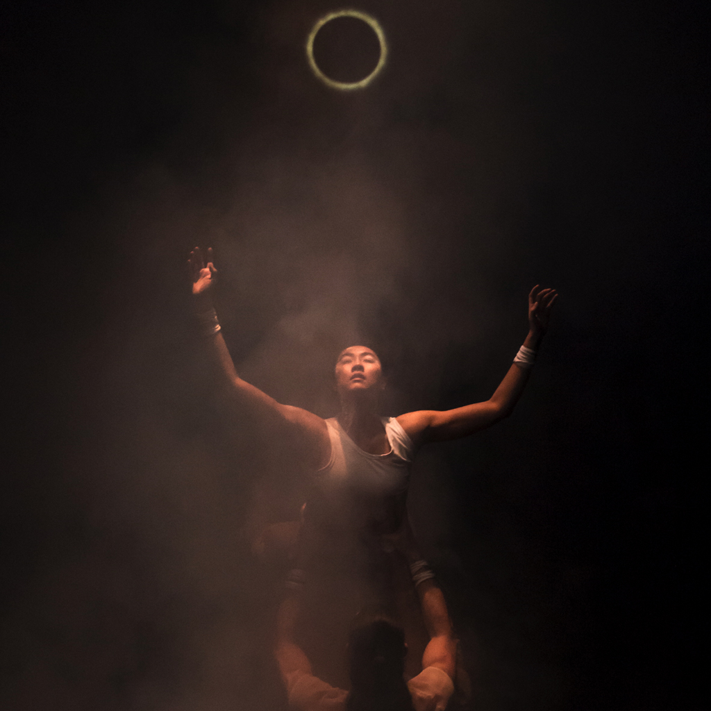 Red Sky Performance: a person in a white tank top arches up towards the sky with their arms spread wide. A person lifts them by the waist underneath. The background is black, and a tan smoke is over the dancers. A ring of light that looks like an eclipsed sun is at the top of the screen.