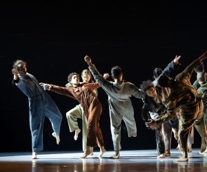 RUBBERBAND: A group of people all falling in a different direction. They are all wearing work jumpsuits of various earth tones. The background is black and there is a spotlight on the performers.