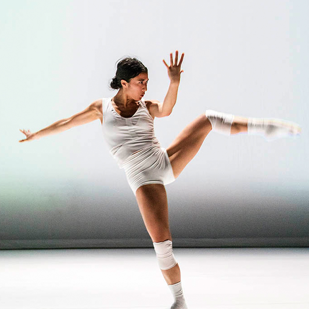Wen Wei Dance: A woman in tight white clothing, socks and knee pads kicks her left leg up and leans back. She looks toward her leg with her hand in front of her eyes. It is a sharp, strong movement. The background and floor is also white.