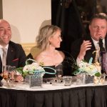 Twisted Black Tie Fundraiser: Andy Fillmore, a white bald man in a black suit sits beside Alicia Orr, a short-haired blonde woman in an off-the-shoulder black dress. To her left is Mayor Mike Savage in a black suit with a purple patterned tie. They are sitting at a table with drinks and white flowers.