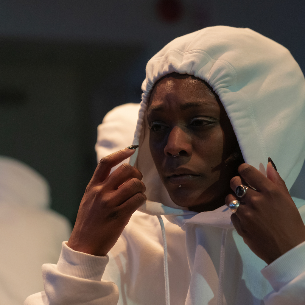 Ebnfloh Dance company: In-ward: a headshot of a dancer in a white hood pulled tight, is pulling on their hood in order to loosen it around their lower face. The dancer's face is making an expression of concern with furrowed brows. They have a nose and lip piercing and black polished nails with two rings on their left hand. The dancer's hands and face contrast against their sweater.